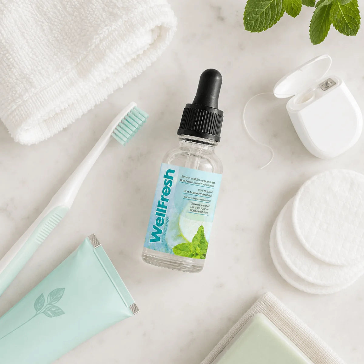 El frasco de WellFresh con gotero sobre mármol, junto a un cepillo de dientes, hilo dental y hojas de menta